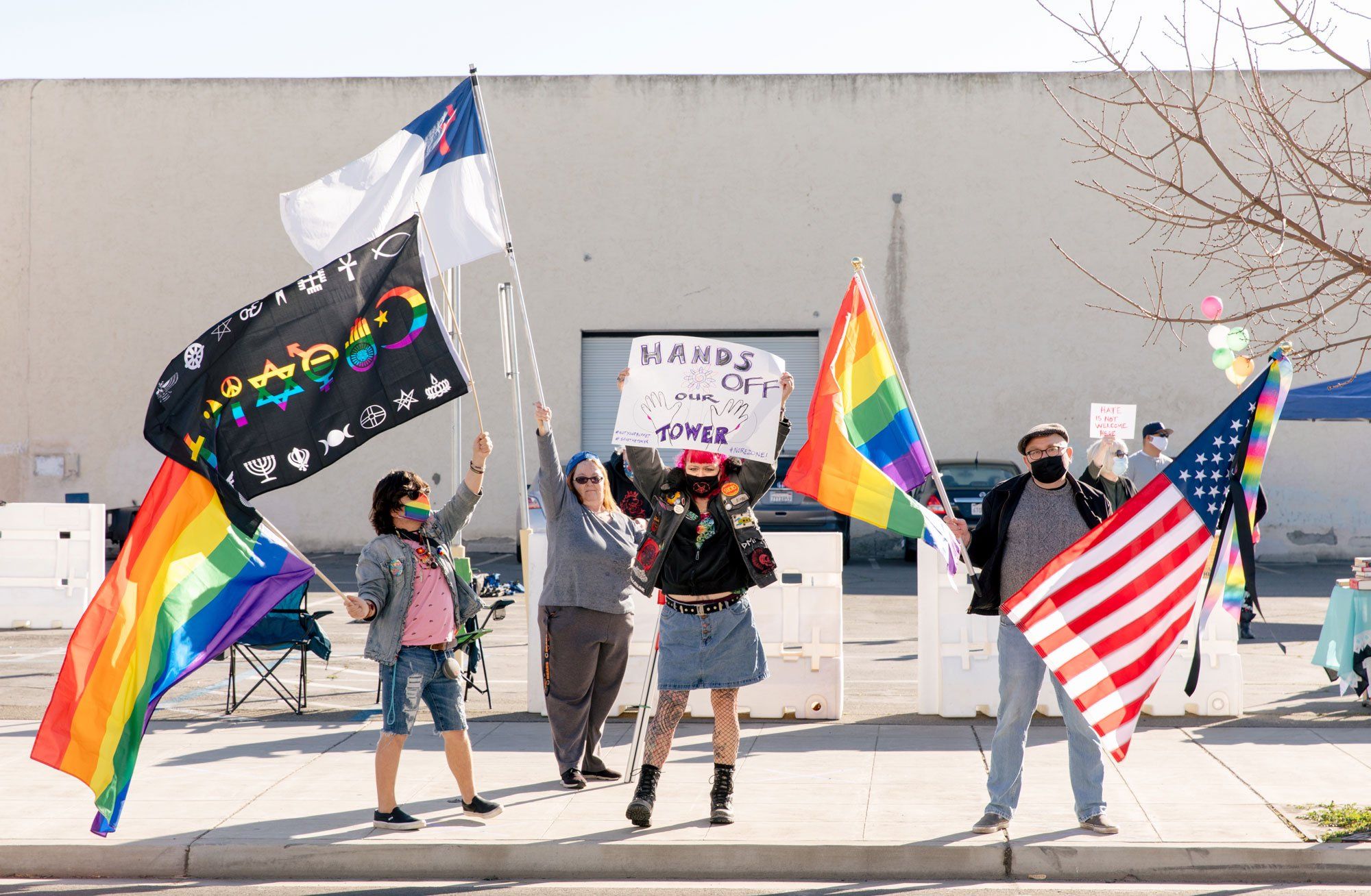 Protesters wave rainbow flags, an American flag and a coexist flag on the sidewalk across the street from the Tower Theatre. A counter protester is in the background waving a white flag containing a red cross.