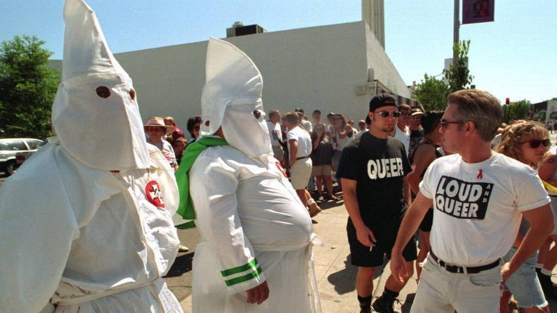 Two individuals dressed in white hoods and robes of the Ku Klux Klan stand at attention among onlookers, two of whomes are wearing shirts with the words “Queer” and “Loud and Queer.”